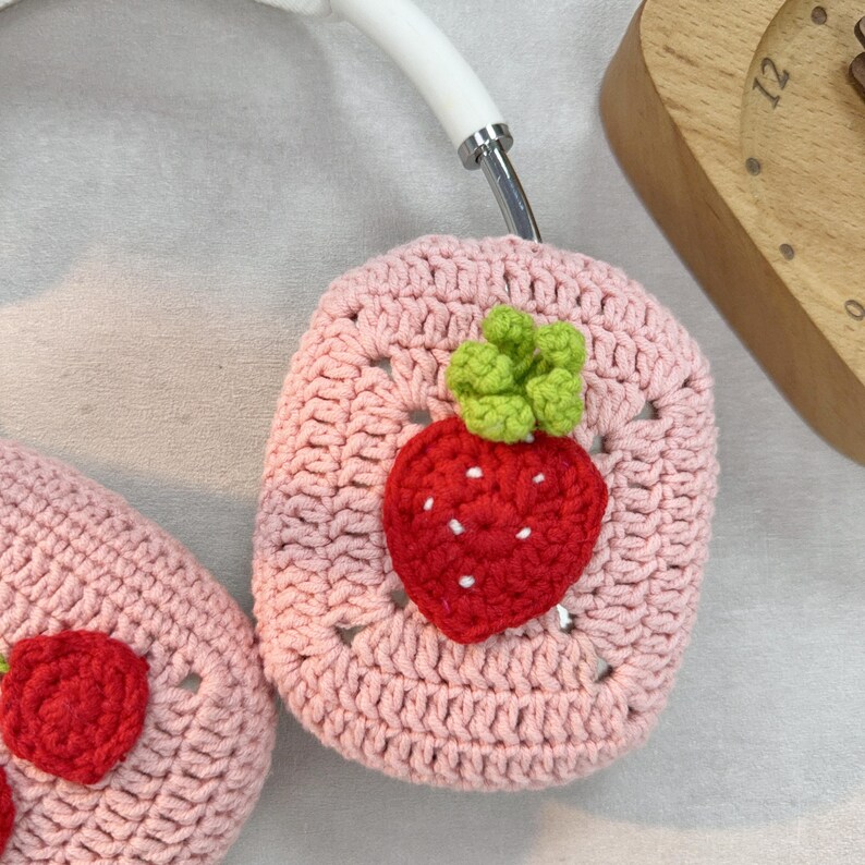 May include: A pair of pink crocheted headphone covers with a strawberry design. The strawberries are red with green leaves.