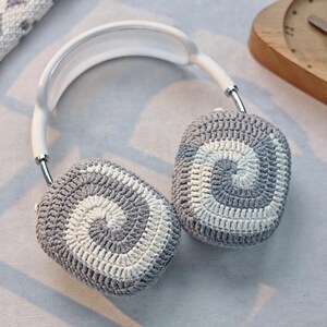 May include: White headphones with grey and white crocheted earmuffs. The earmuffs have a spiral pattern.