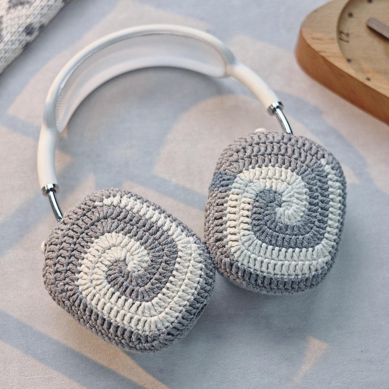 May include: White headphones with grey and white crocheted earmuffs. The earmuffs have a spiral pattern.