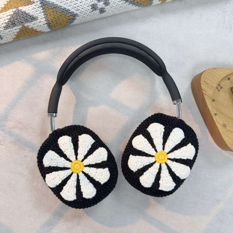 K&ouml;nnte beinhalten: Schwarze Kopfh&ouml;rer mit wei&szlig;en, gestrickten Ohrmuscheln mit G&auml;nsebl&uuml;mchenmuster. Die Ohrmuscheln haben eine gelbe Mitte.