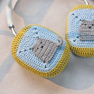 Pu&ograve; includere: Copri cuffie all'uncinetto con un design a gatto grigio. Le cover sono blu e gialle.