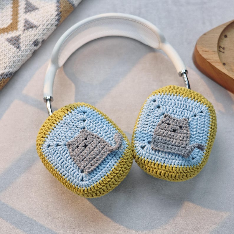 Pu&ograve; includere: Cuffie bianche con paraorecchie all'uncinetto in blu e giallo con un design di gatto grigio.