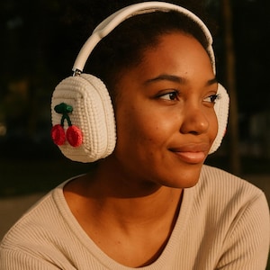 Pode incluir: Fones de ouvido brancos com protetores de ouvido de malha, cada um adornado com um design de cereja vermelha e verde. Os fones de ouvido t&ecirc;m uma faixa de cabe&ccedil;a branca. Uma pessoa usa uma camisa canelada bege.