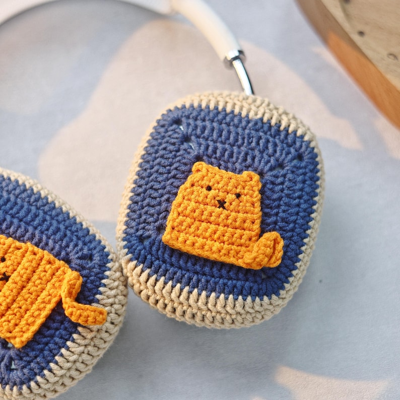 May include: A pair of crocheted headphone covers with a blue and beige background and a yellow cat design.