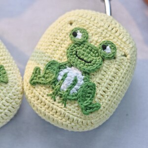 May include: A crocheted yellow and green frog-shaped phone case. The frog has a white belly and black eyes.