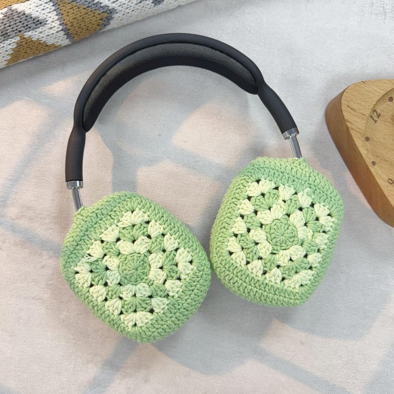 Op de afbeelding: Groene gehaakte oorkappen voor hoofdtelefoons. De oorkappen zijn gemaakt met een granny square-patroon en hebben een lichtgroene en een donkergroene kleur.