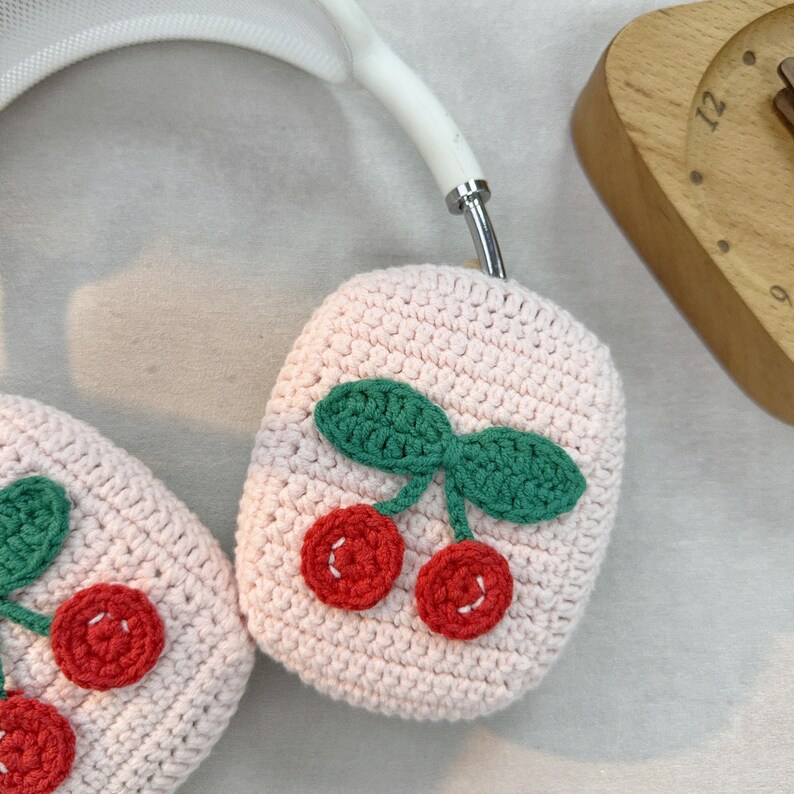 May include: A pair of pink crocheted headphone covers with a cherry design. The cherries are red with green leaves.