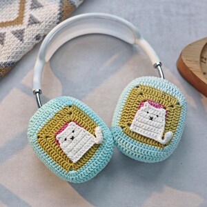 May include: White headphones with crocheted earmuffs featuring a white cat design on a blue and yellow background.