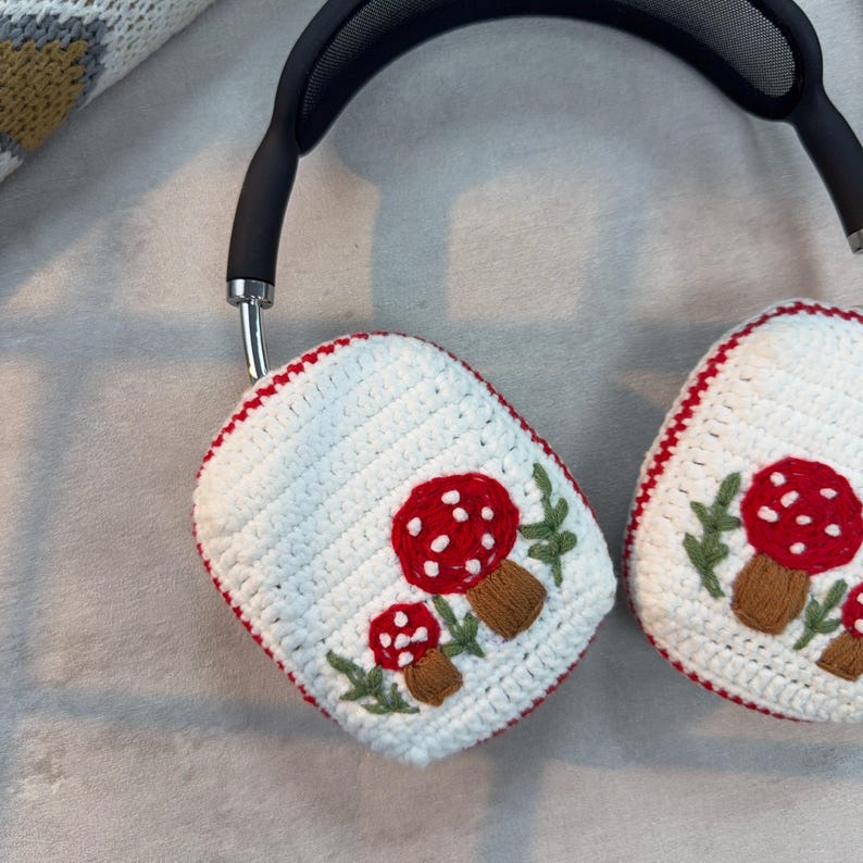 May include: White crocheted headphone covers with red and white mushroom embroidery. The covers are attached to black headphones with a mesh headband.