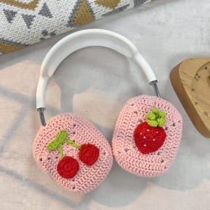 May include: Pink crocheted headphone covers with a strawberry and cherry design. The covers are made with a pink yarn and feature embroidered details of a strawberry with green leaves and a cherry with a green stem.