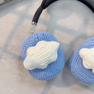 May include: A pair of blue crocheted headphone covers with white crocheted cloud designs. The covers are attached to a black headband.