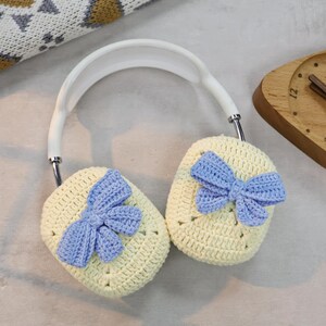 May include: White headphones with crocheted earmuffs. The earmuffs are light yellow with a blue crocheted bow on each side.