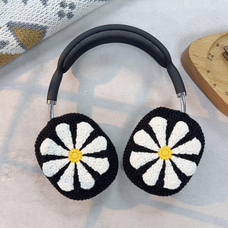 K&ouml;nnte beinhalten: Schwarze Kopfh&ouml;rer mit geh&auml;kelten Ohrensch&uuml;tzern, die ein wei&szlig;es G&auml;nsebl&uuml;mchen-Design mit gelber Mitte zeigen.