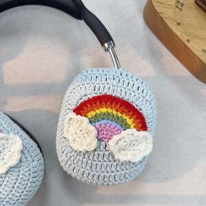 May include: Light blue crochet headphone covers with a colorful rainbow and white cloud accents. The rainbow features red, orange, yellow, green, blue, and purple. The covers are designed to fit over the ear cups of headphones.