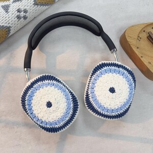 Puede incluir: Un par de orejeras de punto de ganchillo blancas, azules y azul marino para auriculares. Las orejeras son redondas y tienen un centro blanco con un borde azul y azul marino.
