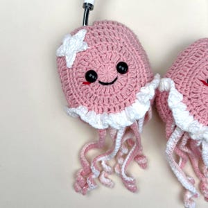 Puede incluir: Medusas rosas hechas a mano con ganchillo y detalles blancos, incluyendo una estrella y un ribete con volantes. Las medusas tienen ojos negros y una cara sonriente. Los tent&aacute;culos son rosas y blancos. Dise&ntilde;o caprichoso.