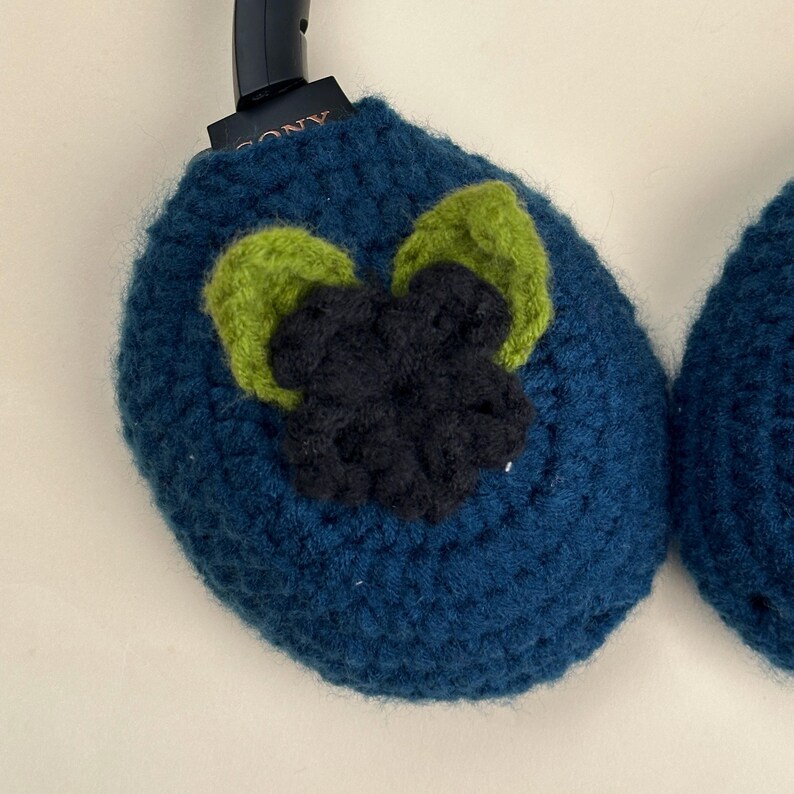May include: A pair of blue crochet headphone covers with a green and black floral design. The covers are designed to fit over the earcups of a pair of headphones. The Sony logo is visible on the headphones.