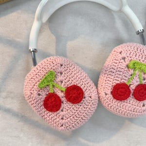 K&ouml;nnte beinhalten: Rosa gestrickte Kopfh&ouml;rerabdeckungen mit einem Kirschendesign. Die Abdeckungen sind aus weichem Garn gefertigt und zeigen ein Paar rote Kirschen mit gr&uuml;nen Stielen und Bl&auml;ttern.