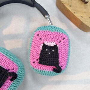 May include: A pair of pink and teal crocheted headphone covers with a black crocheted cat design.