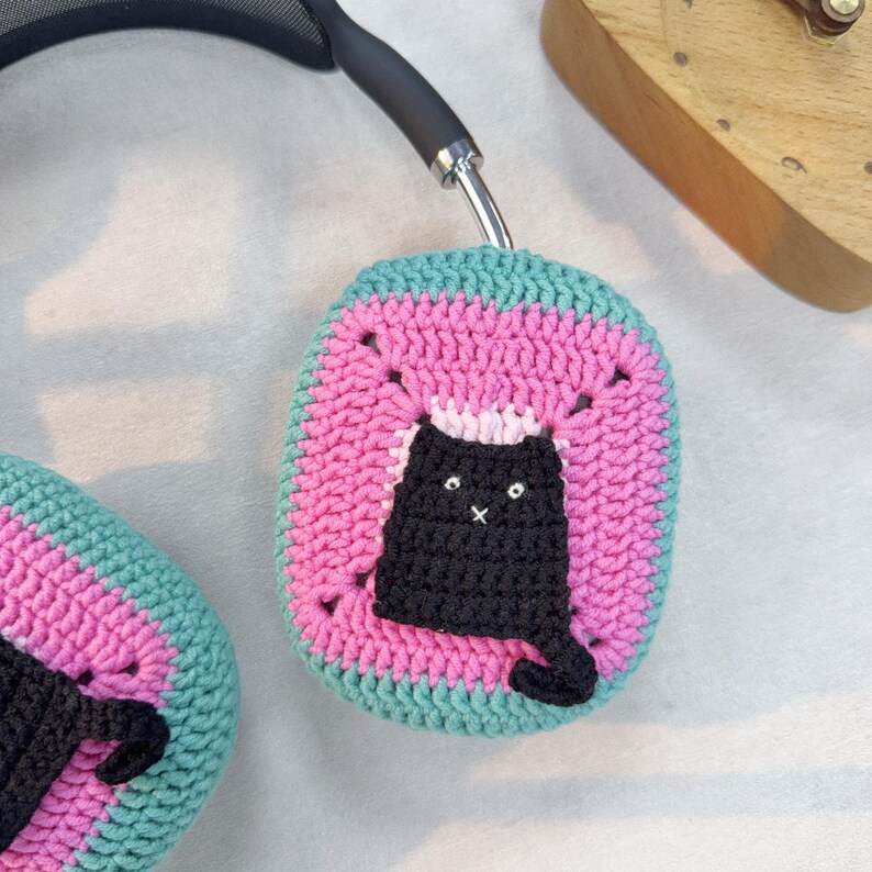 May include: A pair of pink and teal crocheted headphone covers with a black crocheted cat design.