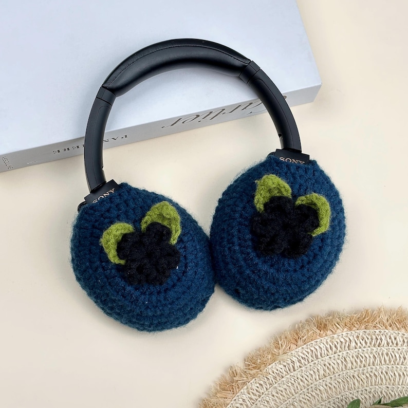 May include: Blue crochet ear covers with black and green floral accents, attached to black headphones. The ear covers are a dark blue color, with a black flower and green leaves design. The headphones have the brand name "SONY" printed on them.
