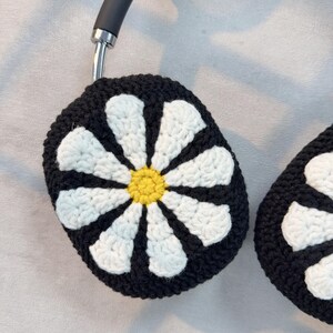 K&ouml;nnte beinhalten: Schwarze gestrickte Kopfh&ouml;rerabdeckungen mit einem wei&szlig;en G&auml;nsebl&uuml;mchen-Design. Die G&auml;nsebl&uuml;mchen haben einen gelben Mittelpunkt.