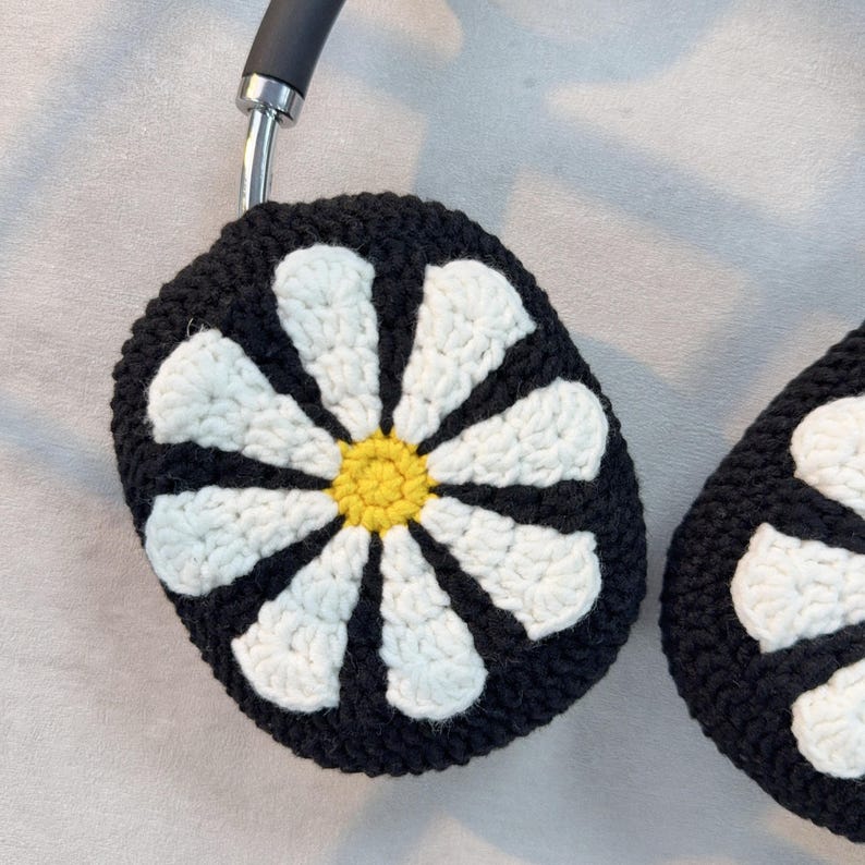 K&ouml;nnte beinhalten: Schwarze gestrickte Kopfh&ouml;rerabdeckungen mit einem wei&szlig;en G&auml;nsebl&uuml;mchen-Design. Die G&auml;nsebl&uuml;mchen haben einen gelben Mittelpunkt.