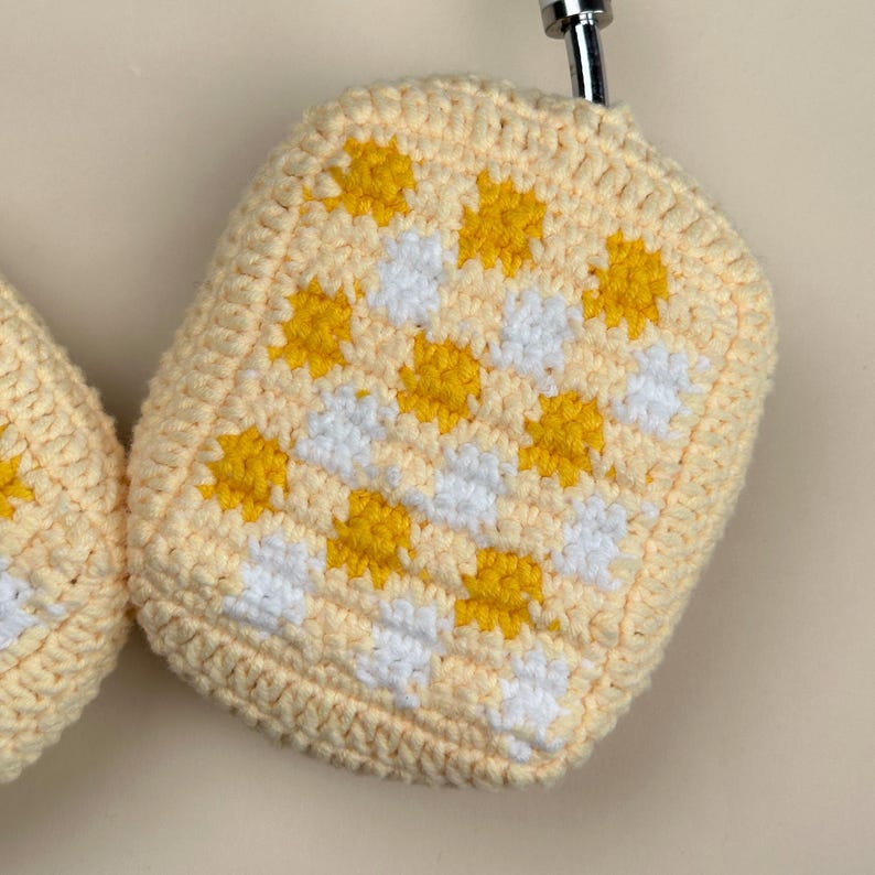 May include: Close-up of a pair of crochet headphone covers. The covers are a light yellow color with a checkerboard pattern of white and yellow squares. The covers are hanging from a silver hook.