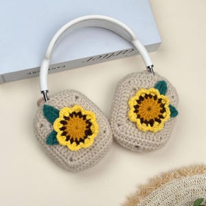 Op de afbeelding: Cr&egrave;mekleurige koptelefoon met gehaakte oorschelpen. Elke hoes heeft een gele en bruine zonnebloem met groene bladeren. De hoofdband is wit en de oorschelpen zijn vierkant.