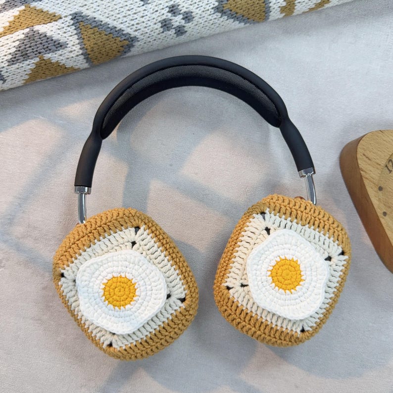 May include: A pair of headphones with crocheted earmuffs. The earmuffs are brown and white with a yellow circle in the center, resembling a fried egg.