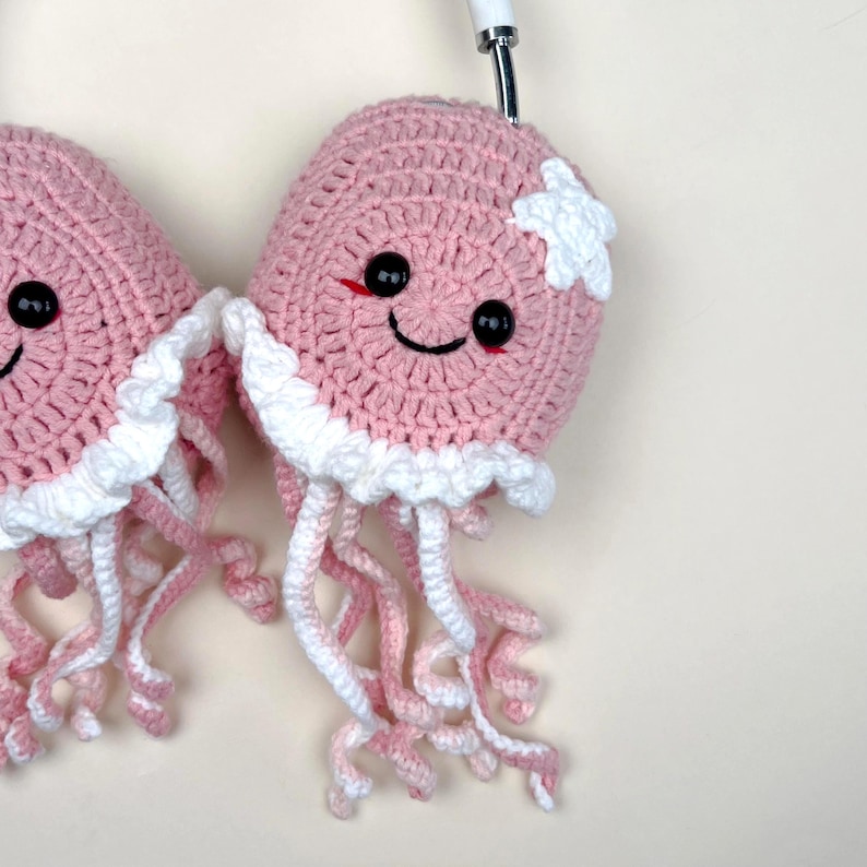 Puede incluir: Dos medusas rosas de ganchillo con tent&aacute;culos blancos y rosas, ojos negros y bocas sonrientes. Una medusa tiene un acento en forma de estrella blanca. Las medusas est&aacute;n unidas a unos auriculares blancos y plateados.