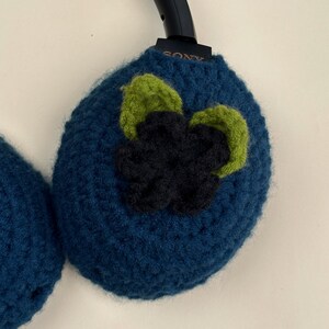 May include: Hand-crocheted headphone covers in a deep teal blue. Each cover is adorned with a three-dimensional flower design in shades of green and navy blue. The Sony logo is visible on the headphone band. These covers add a unique, handmade touch to your headphones.
