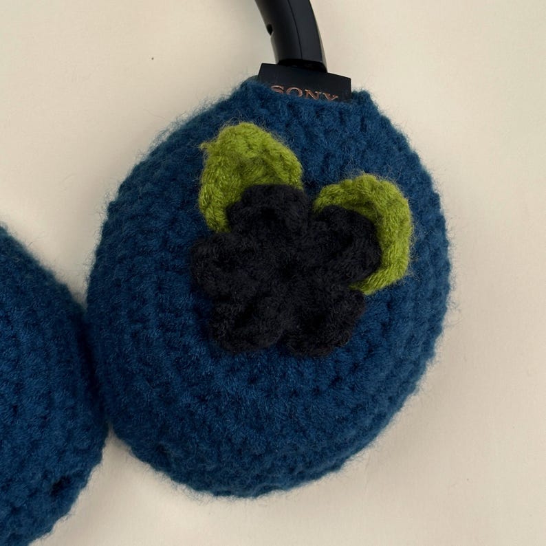 May include: Hand-crocheted headphone covers in a deep teal blue. Each cover is adorned with a three-dimensional flower design in shades of green and navy blue. The Sony logo is visible on the headphone band. These covers add a unique, handmade touch to your headphones.