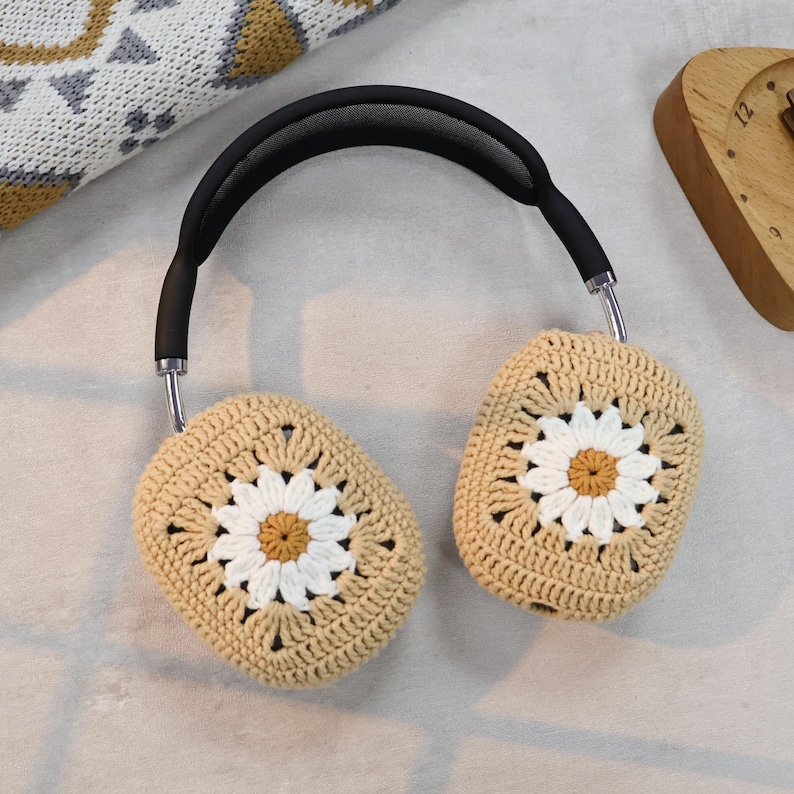 Puede incluir: Fundas para auriculares de crochet beige con un dise&ntilde;o de margarita blanca. Las fundas est&aacute;n unidas a una diadema negra.