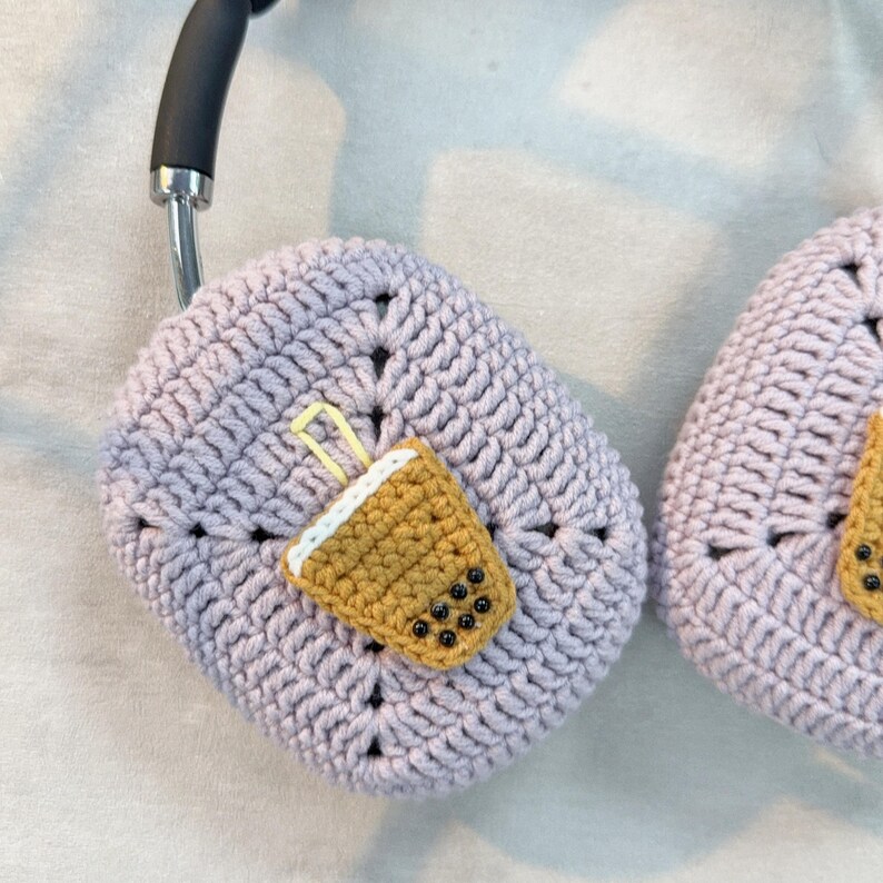 May include: A pair of crocheted headphone covers in a light purple colour. One cover features a crocheted brown and yellow boba tea design with black boba pearls.