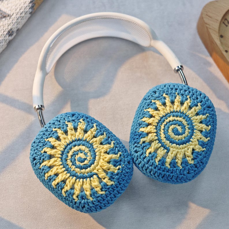 May include: White headphones with blue and yellow crocheted earmuffs featuring a sun design.