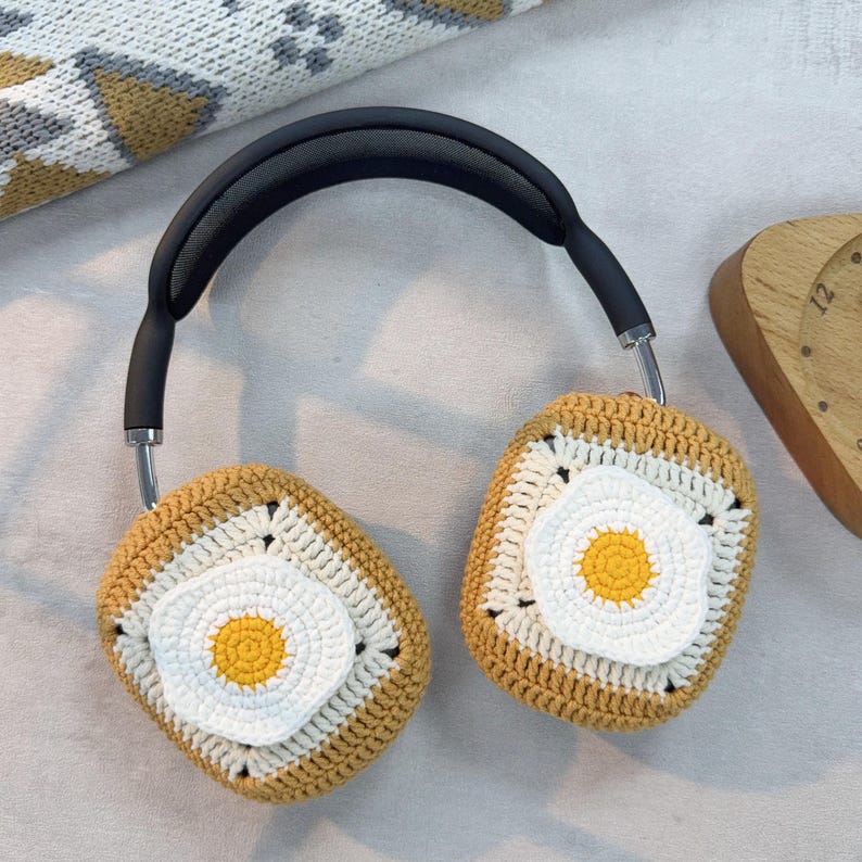 May include: Crocheted headphone covers in the shape of a fried egg. The covers are brown and white with a yellow yolk in the center. The headphones are black with a silver headband.