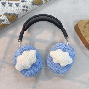 May include: Blue knitted earmuffs with white cloud accents. The earmuffs are attached to a black headband.