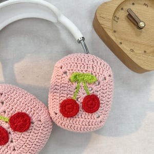K&ouml;nnte beinhalten: Rosa gestrickte Kopfh&ouml;rerabdeckungen mit einem Kirschendesign. Die Kirschen sind rot mit gr&uuml;nen Stielen und Bl&auml;ttern.