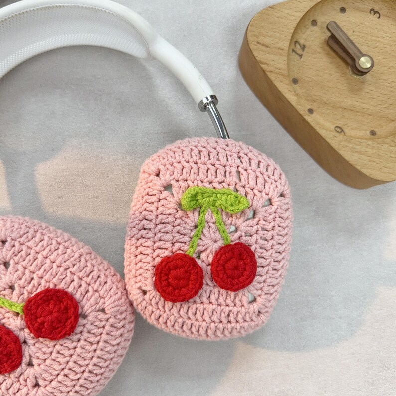 K&ouml;nnte beinhalten: Rosa gestrickte Kopfh&ouml;rerabdeckungen mit einem Kirschendesign. Die Kirschen sind rot mit gr&uuml;nen Stielen und Bl&auml;ttern.