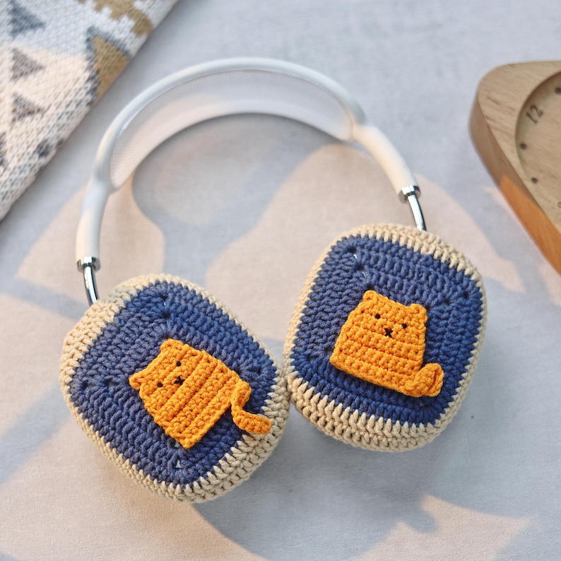 May include: White headphones with crocheted earmuffs featuring a yellow cat design. The earmuffs are blue and beige.
