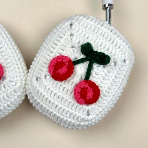 Pode incluir: Capas de fone de ouvido de croch&ecirc; branco com um design de cereja vermelha e rosa. As cerejas t&ecirc;m caules e folhas verdes. As capas s&atilde;o quadradas e t&ecirc;m uma apar&ecirc;ncia texturizada, com um padr&atilde;o de ponto vis&iacute;vel.