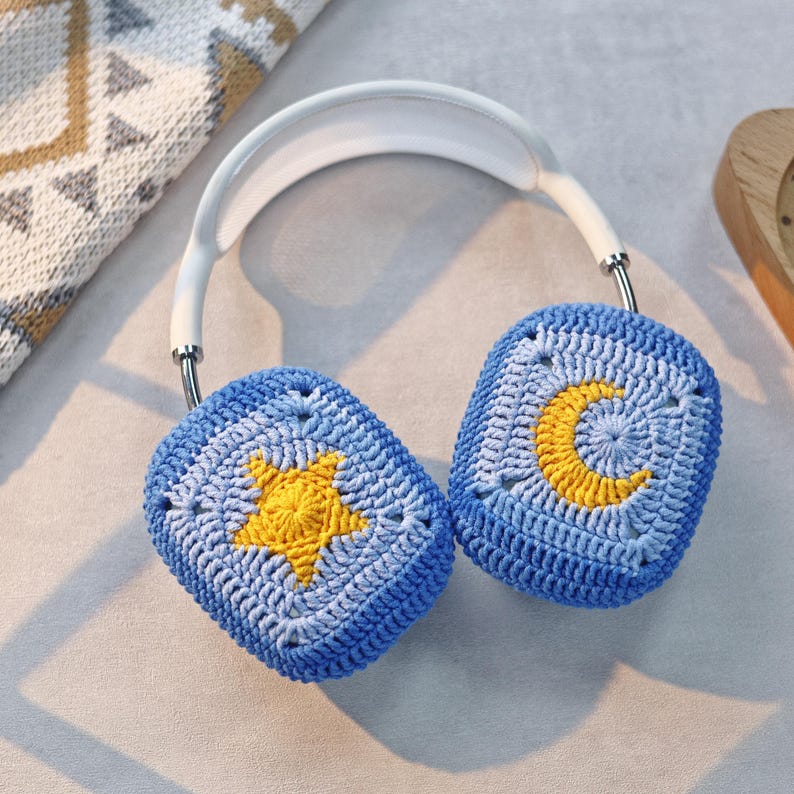 Puede incluir: Cubiertas para auriculares de crochet azul con un dise&ntilde;o de estrella amarilla y luna creciente.