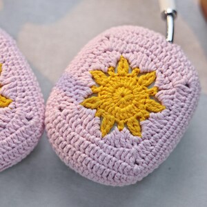 K&ouml;nnte beinhalten: Zwei rosa geh&auml;kelte Kopfh&ouml;rerh&uuml;llen mit einem gelben geh&auml;kelten Sonnendesign.