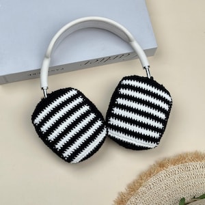 May include: Headphones with white headband and black and white striped ear covers. The ear covers are crocheted with horizontal black and white stripes. The headphones are on a light beige surface.