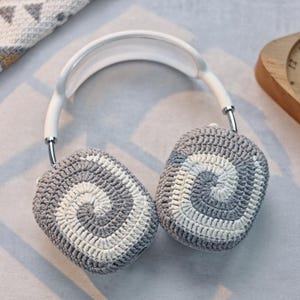 May include: White headphones with grey and white crocheted ear covers. The ear covers have a spiral pattern.