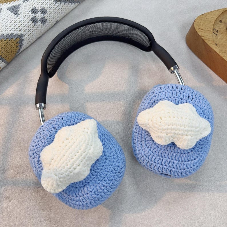May include: Blue crocheted headphone covers with white crocheted cloud designs. The covers are attached to a black headband with a mesh design.