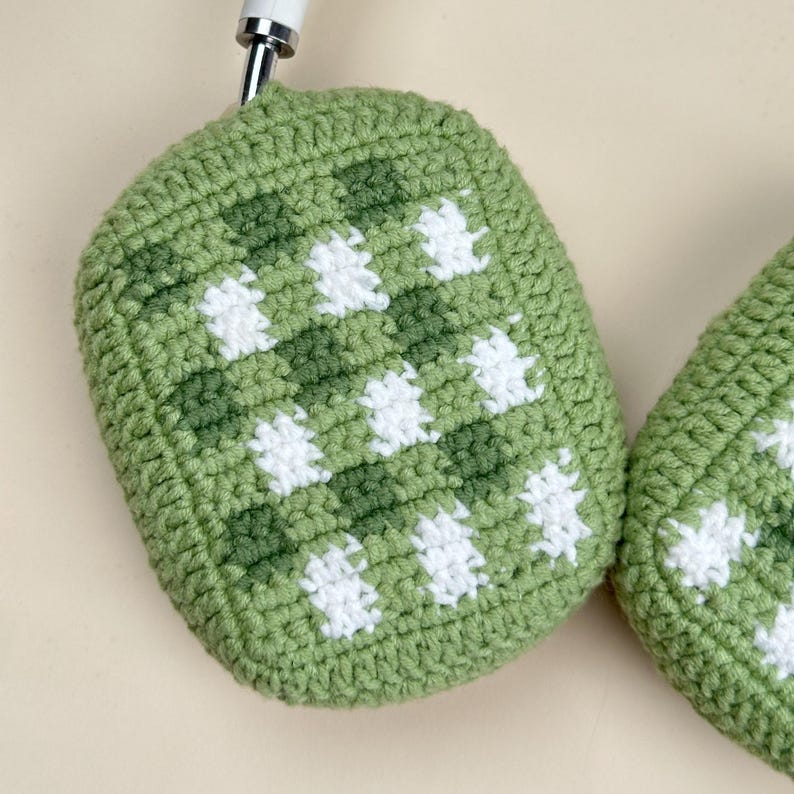 May include: Close-up of a green and white crocheted headphone cover. The cover has a checkered pattern with alternating squares of light and dark green and white. The headphones are partially visible, with a white and silver handle.