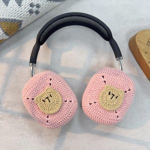 May include: Pink crocheted headphone covers with a brown bear face design. The covers are made of soft yarn and are designed to fit over the earcups of headphones.
