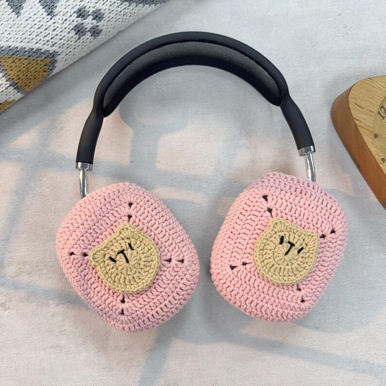 May include: Pink crocheted headphone covers with a brown bear face design. The covers are made of soft yarn and are designed to fit over the earcups of headphones.